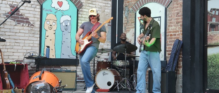 A three-person band performing on an outdoor stage: two guitarists and a drummer, with a brick wall and cartoon llama artwork in the background.